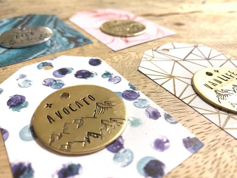 May include: Close-up of several small, round, gold-colored metal tags with various designs and text, including "AVOCATO" and "VANLIFE". The tags are attached to decorative paper tags with patterns in blue, pink, and gold.