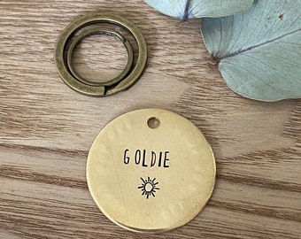 May include: Brass dog tag with the name "Goldie" and a sun graphic engraved on it. The tag is attached to a split ring.