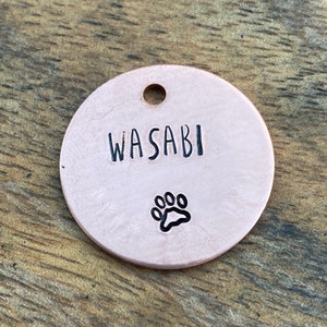 May include: A round, light pink metal dog tag with the name "WASABI" engraved in black lettering and a black paw print.