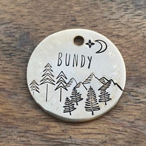 May include: A round, gold-colored metal dog tag with a mountain and forest scene engraved on it. The tag also has the name "BUNDY" engraved on it, along with a star and crescent moon.