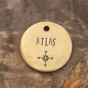 Könnte beinhalten: Ein runder Messinganhänger mit dem Wort "ATLAS" eingraviert. Unter dem Wort ist eine Kompassrose mit den Richtungen N, S, E und W eingraviert.
