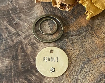 May include: A round, brass dog tag with the name "Peanut" engraved on it. A small paw print is engraved below the name. The tag is attached to a split ring.