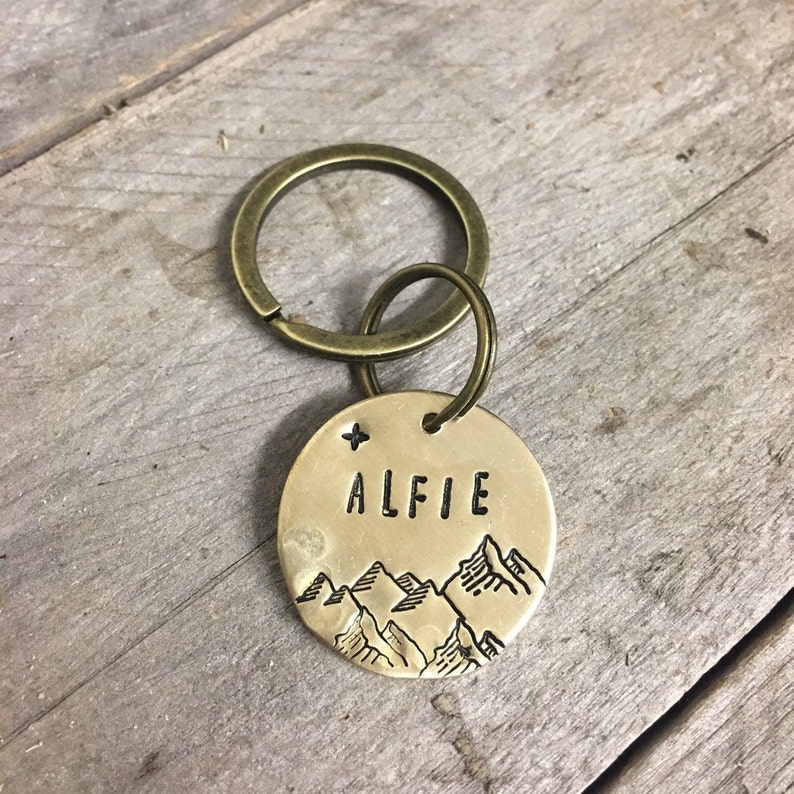 May include: A round, brass-colored dog tag with the name "ALFIE" stamped in block letters. The tag features a mountain range design and a star. Attached is a brass-colored split ring and a larger ring.