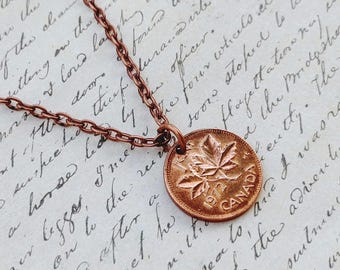Pode incluir: Um colar de corrente de metal em tom de cobre com um pingente circular feito de uma moeda canadense de um centavo de 1972. A moeda exibe um design de folha de bordo, o texto 1972 e a palavra CANADA, suspensa por uma corrente de elos.