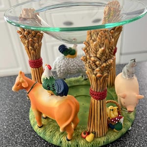 May include: A decorative farm-themed oil warmer with a clear glass dish. The base features a cow, pig, sheep, and rooster figurines. The warmer has wheat stalk supports tied with red twine and a basket of fruit.