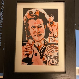 May include: Framed watercolor painting of a woman with dark hair and a pink shirt, looking directly at the viewer. A man with a light orange shirt is standing behind her. The painting is set against a dark background.