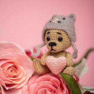May include: A crocheted teddy bear wearing a gray cat-eared hat holds a pink heart in its paws. The bear is sitting in a bouquet of pink roses.
