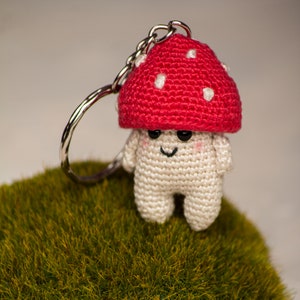 May include: A crocheted mushroom keychain with a red cap and white spots. The mushroom has a white body with a smiling face and black eyes. The keychain is attached to a silver metal ring.