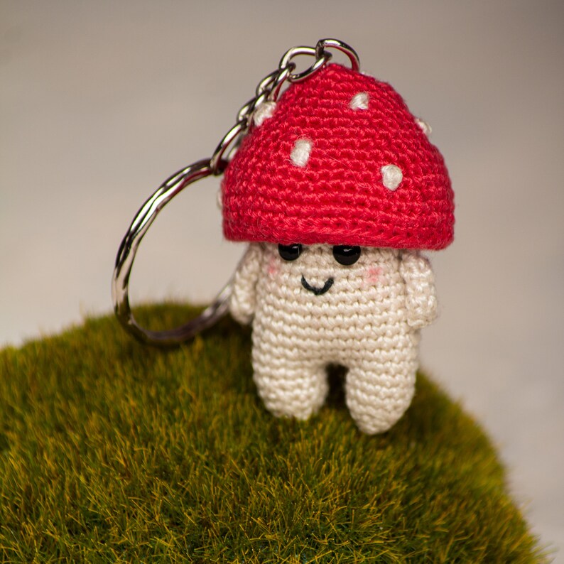 May include: A crocheted mushroom keychain with a red cap and white spots. The mushroom has a white body with a smiling face and black eyes. The keychain is attached to a silver metal ring.