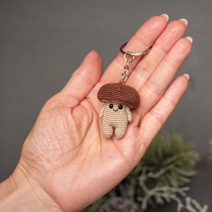 May include: A brown and beige crocheted mushroom keychain with a smiling face. The keychain is attached to a silver metal ring.