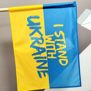 May include: A blue and yellow flag with the text "I Stand With Ukraine" on the blue side and "Ukraine" on the yellow side.