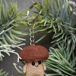 May include: A crocheted mushroom keychain with a brown cap and a beige body. The mushroom has a smiling face with black eyes. The keychain is attached to a silver key ring.