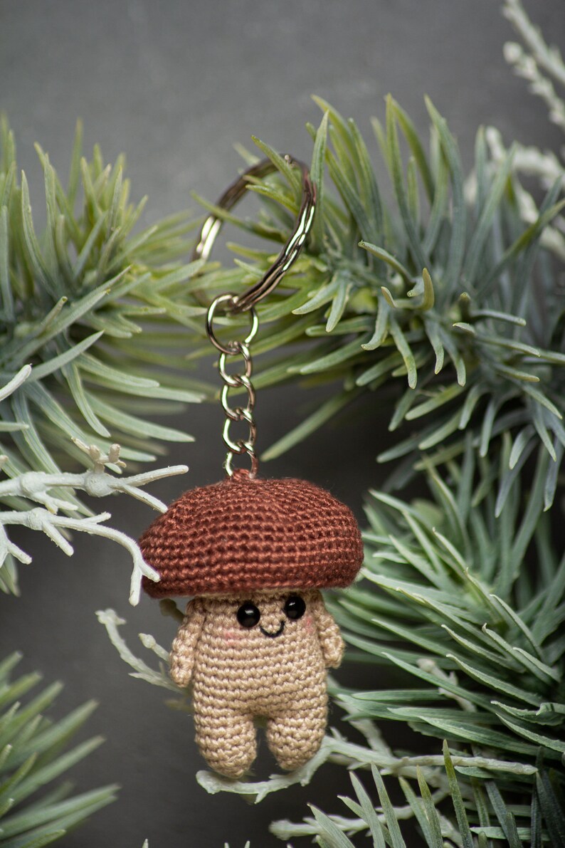 May include: A crocheted mushroom keychain with a brown cap and a beige body. The mushroom has a smiling face with black eyes. The keychain is attached to a silver key ring.
