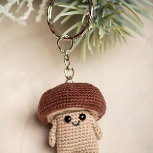 May include: A crocheted mushroom keychain with a brown cap and a beige body. The mushroom has a smiling face with black eyes and a small mouth. The keychain is attached to a silver key ring.