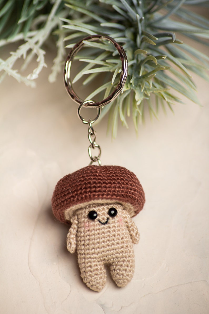 May include: A crocheted mushroom keychain with a brown cap and a beige body. The mushroom has a smiling face with black eyes and a small mouth. The keychain is attached to a silver key ring.