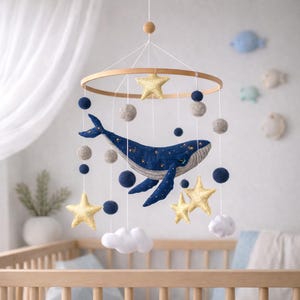 Può includere: Mobile per bambini con una balena blu navy e dettagli di stelle dorate, sospeso a un cerchio di legno. Il mobile include stelle dorate, nuvole bianche e palline di feltro blu navy e grigie, creando una decorazione per la cameretta.