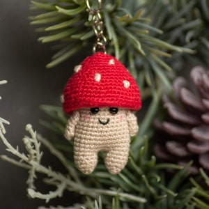 May include: A crocheted mushroom keychain with a red cap and white spots. The mushroom has a smiling face and is hanging from a silver chain.