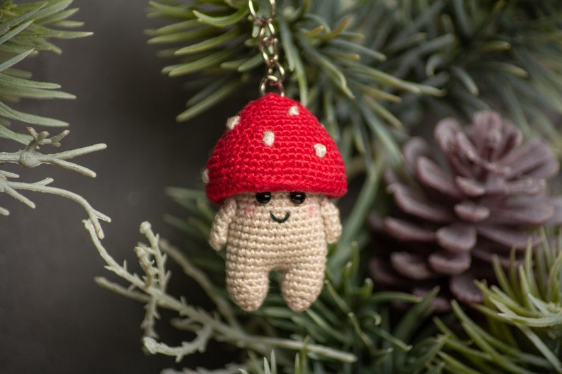 May include: A crocheted mushroom keychain with a red cap and white spots. The mushroom has a smiling face and is hanging from a silver chain.