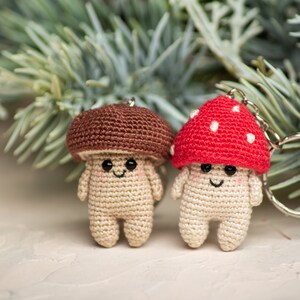 May include: Two crocheted mushroom keychains, one brown and one red with white dots. The keychains are smiling and have black eyes. They are hanging from a silver keychain ring.