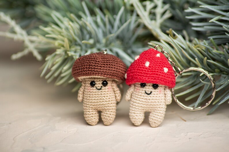 May include: Two crocheted mushroom keychains, one brown and one red with white dots. The keychains are smiling and have black eyes. They are hanging from a silver keychain ring.