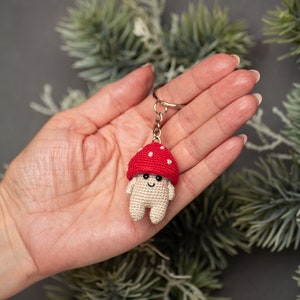 May include: A crocheted mushroom keychain with a red cap and white spots. The mushroom has a smiling face and small white arms and legs.