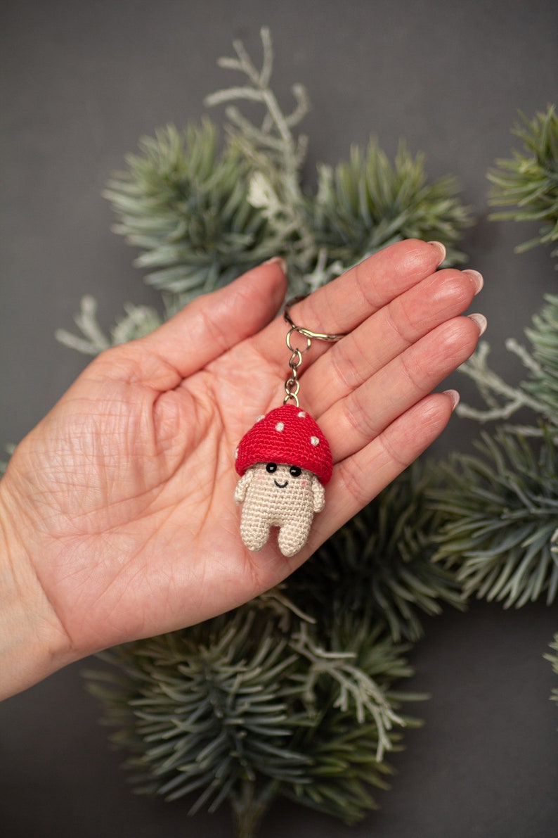 May include: A crocheted mushroom keychain with a red cap and white spots. The mushroom has a smiling face and small white arms and legs.