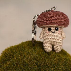 May include: A crocheted mushroom keychain with a brown cap and a beige body. The mushroom has a smiling face with black eyes and a small mouth. The keychain is attached to a silver key ring.