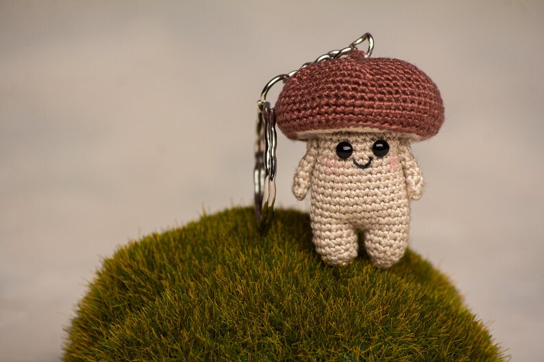 May include: A crocheted mushroom keychain with a brown cap and a beige body. The mushroom has a smiling face with black eyes and a small mouth. The keychain is attached to a silver key ring.