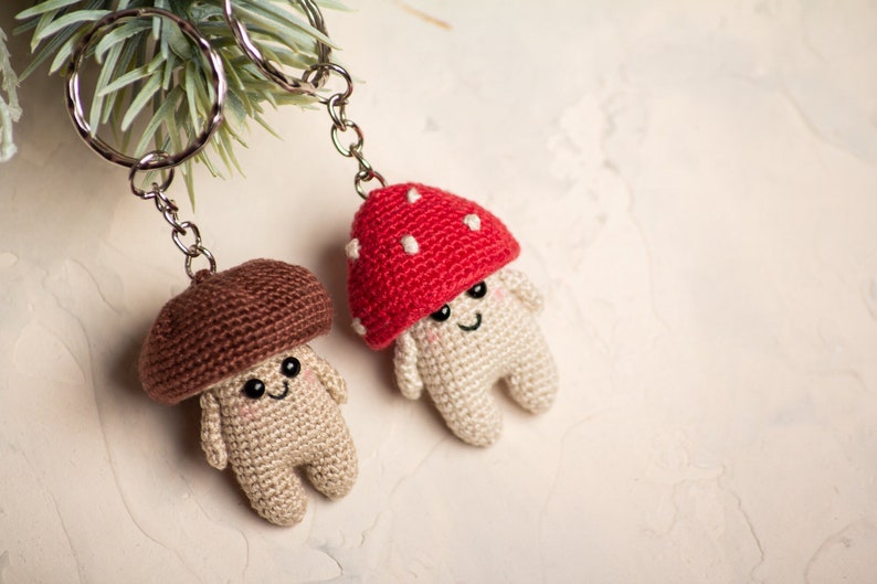 May include: Two crocheted mushroom keychains with smiling faces. One mushroom is brown and the other is red with white dots.