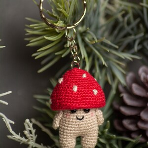 May include: A crocheted mushroom keychain with a red cap and white spots. The mushroom has a smiling face and is attached to a silver key ring.
