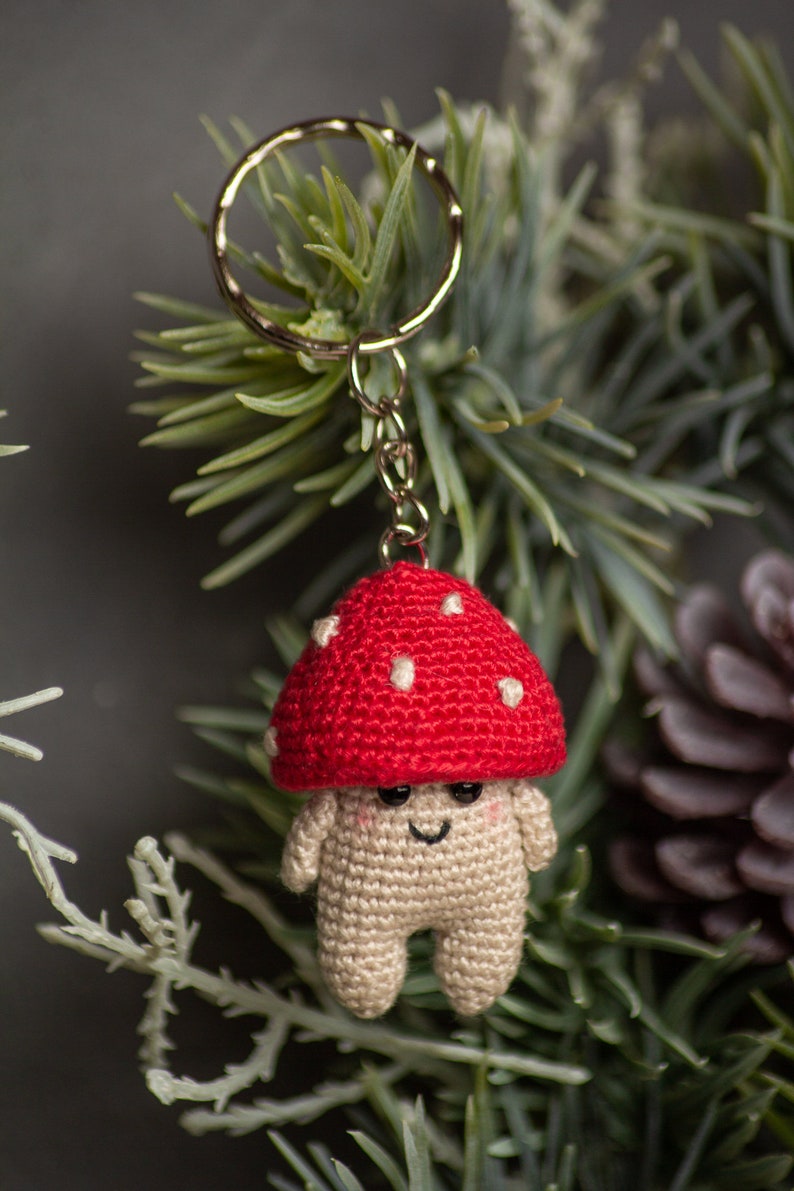 May include: A crocheted mushroom keychain with a red cap and white spots. The mushroom has a smiling face and is attached to a silver key ring.
