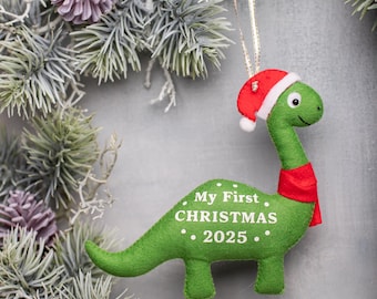 May include: A green dinosaur Christmas ornament with a red Santa hat and scarf. The ornament has the text "My First Christmas • 2025." The ornament is hanging from a gold string, and is surrounded by evergreen branches and pinecones.