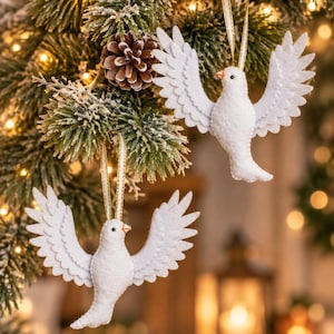 May include: Two white dove ornaments crafted from felt, each featuring spread wings, a small orange beak, and a black eye. Each bird is suspended by a thin gold ribbon loop, designed for hanging display with detailed stitching along the wing edges.
