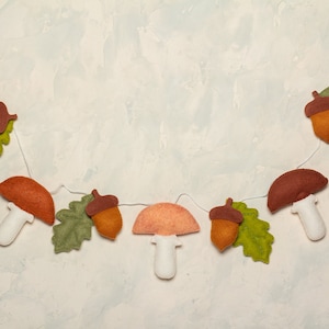 May include: A felt garland with mushrooms, acorns, and leaves in shades of brown, green, and orange. The garland is strung on a white cord and is perfect for fall decor.