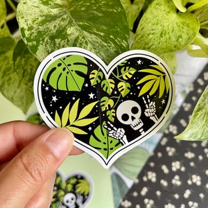 May include: A heart-shaped sticker with a black background and a skeleton with peace signs. The sticker features green and yellow tropical leaves and white stars.