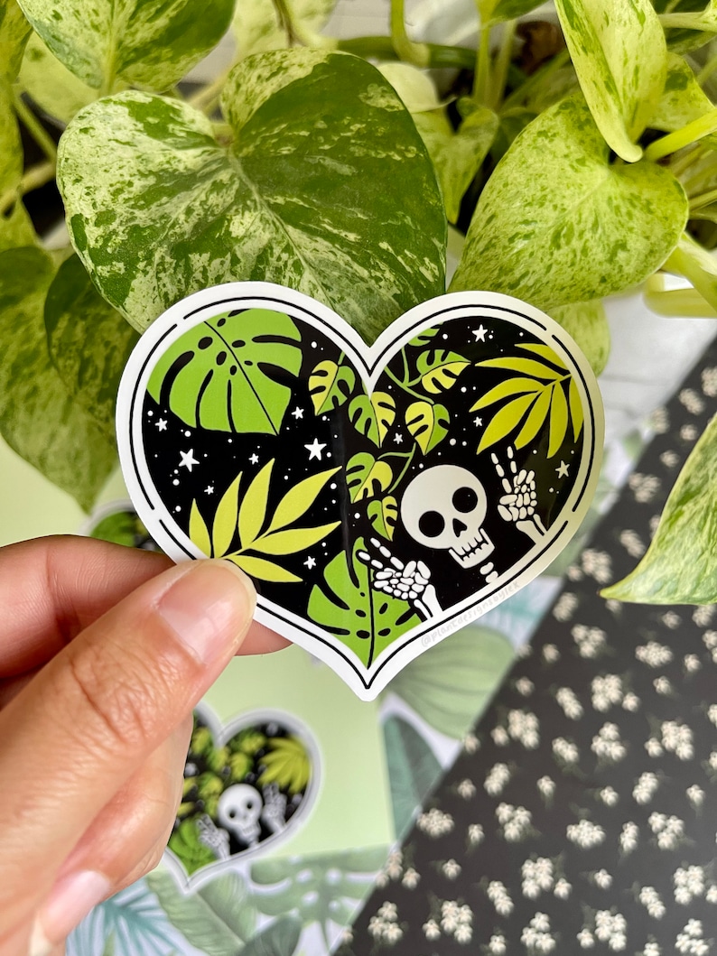 May include: A heart-shaped sticker with a black background and a skeleton with peace signs. The sticker features green and yellow tropical leaves and white stars.