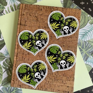 May include: Four heart-shaped stickers with a black background, green leaves, white stars, and a white skeleton with a peace sign. The stickers are on a corkboard.