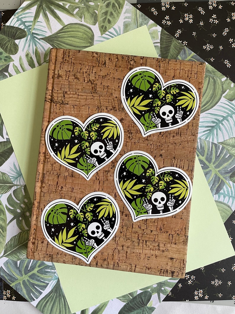 May include: Four heart-shaped stickers with a black background, green leaves, white stars, and a white skeleton with a peace sign. The stickers are on a corkboard.