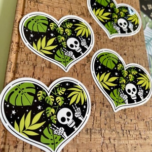 May include: Four heart-shaped stickers with a black background, white stars, and green tropical leaves. Each sticker features a white skeleton with a skull, making a peace sign with its hands.