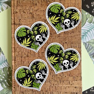 May include: Four heart-shaped stickers with a black background and green tropical leaves. Each sticker features a white skeleton with a skull wearing a peace sign.