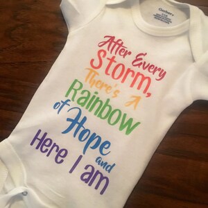 May include: White baby onesie with a rainbow-colored graphic that reads "After Every Storm, There's A Rainbow of Hope and Here I am".
