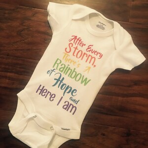 May include: White baby onesie with a rainbow-colored graphic that reads "After Every Storm, There's A Rainbow of Hope and Here I am".