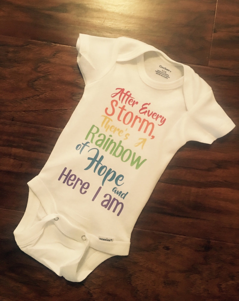 May include: White baby onesie with a rainbow-colored graphic that reads "After Every Storm, There's A Rainbow of Hope and Here I am".