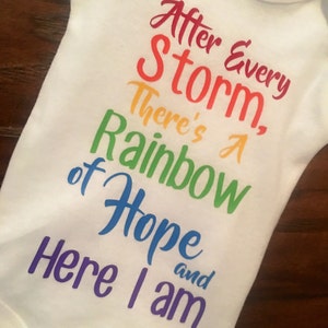 May include: White baby onesie with a rainbow-colored graphic that reads "After Every Storm, There's A Rainbow of Hope and Here I am".