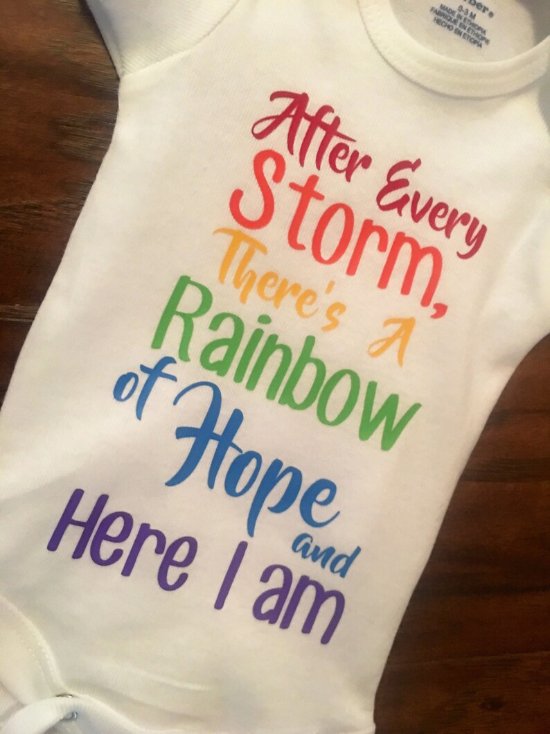 May include: White baby onesie with a rainbow-colored graphic that reads "After Every Storm, There's A Rainbow of Hope and Here I am".