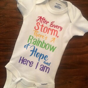 May include: White baby onesie with a rainbow-colored graphic that reads "After Every Storm, There's A Rainbow of Hope and Here I am."