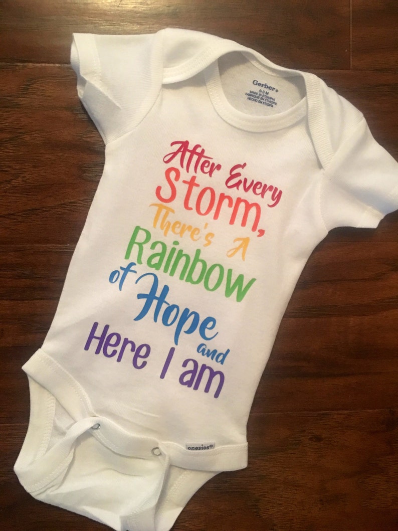 May include: White baby onesie with a rainbow-colored graphic that reads "After Every Storm, There's A Rainbow of Hope and Here I am."