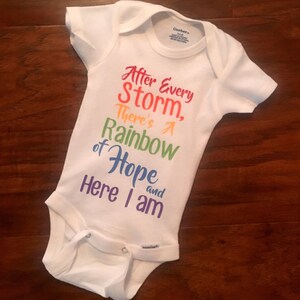 May include: White baby onesie with the text "After Every Storm, There's A Rainbow of Hope and Here I Am" printed in rainbow colors.