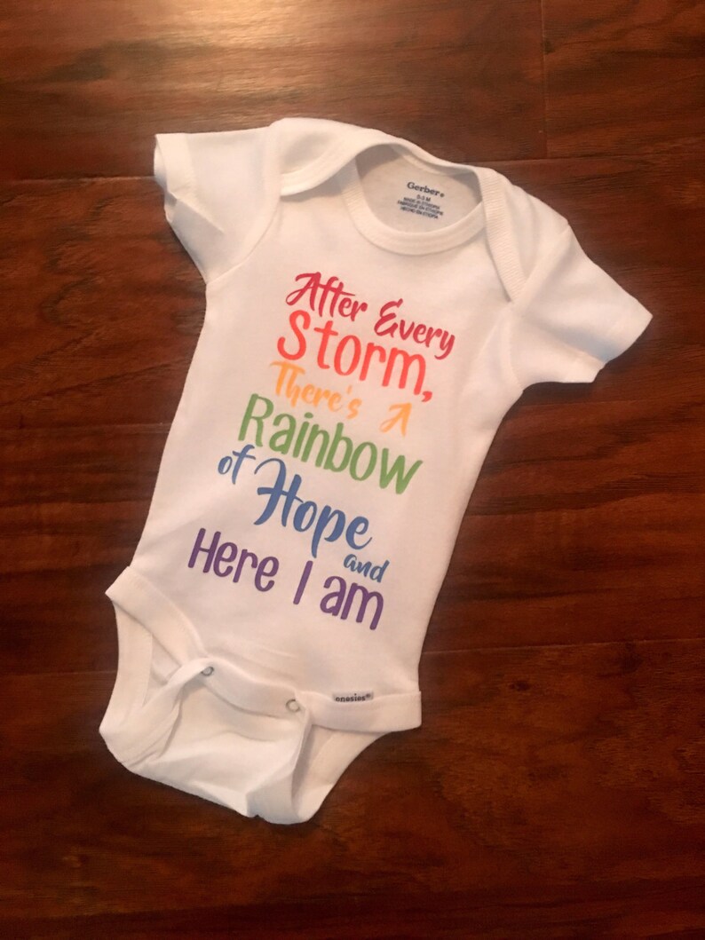 May include: White baby onesie with the text "After Every Storm, There's A Rainbow of Hope and Here I Am" printed in rainbow colors.