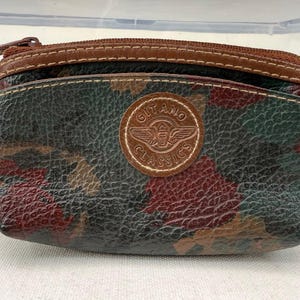 May include: A small, textured coin purse featuring a camouflage pattern in shades of dark green, brown, and red. The purse has a brown leather zipper trim and a circular brown leather patch in the center embossed with the text Gitano Classics.